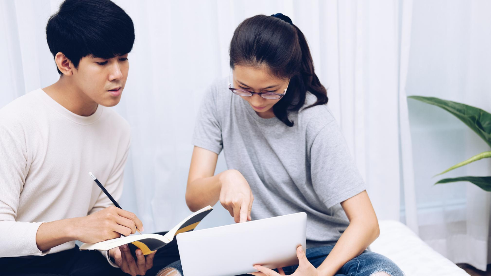 man and a woman sit at their couch, focused on their screens as they take an online course and make notes