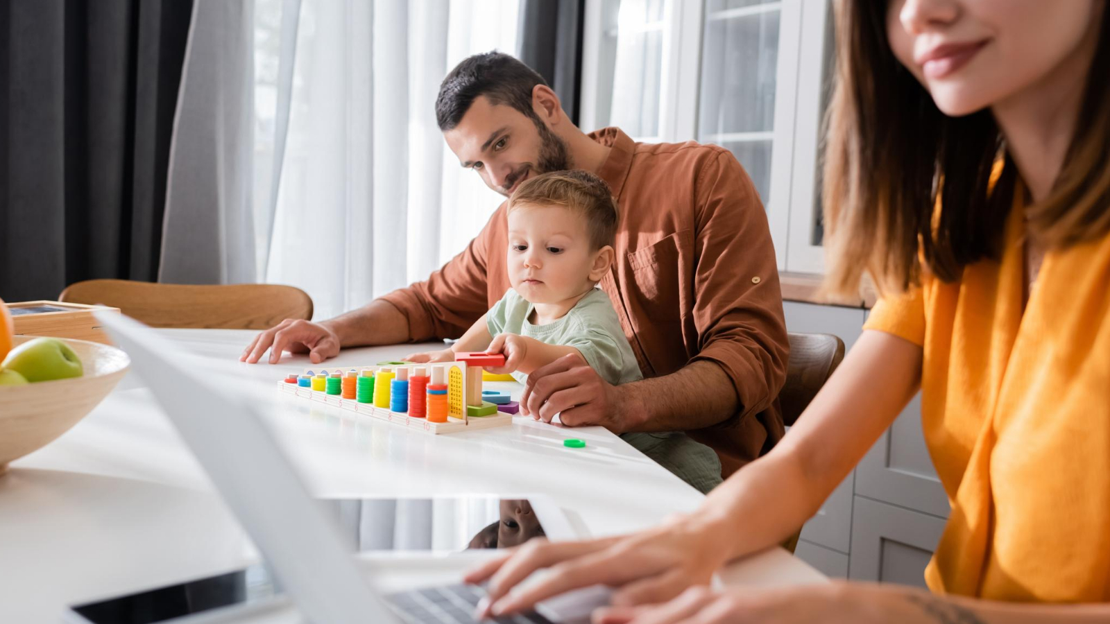 woman sits at her home office desk, working on her laptop as her husband and kid play beside her